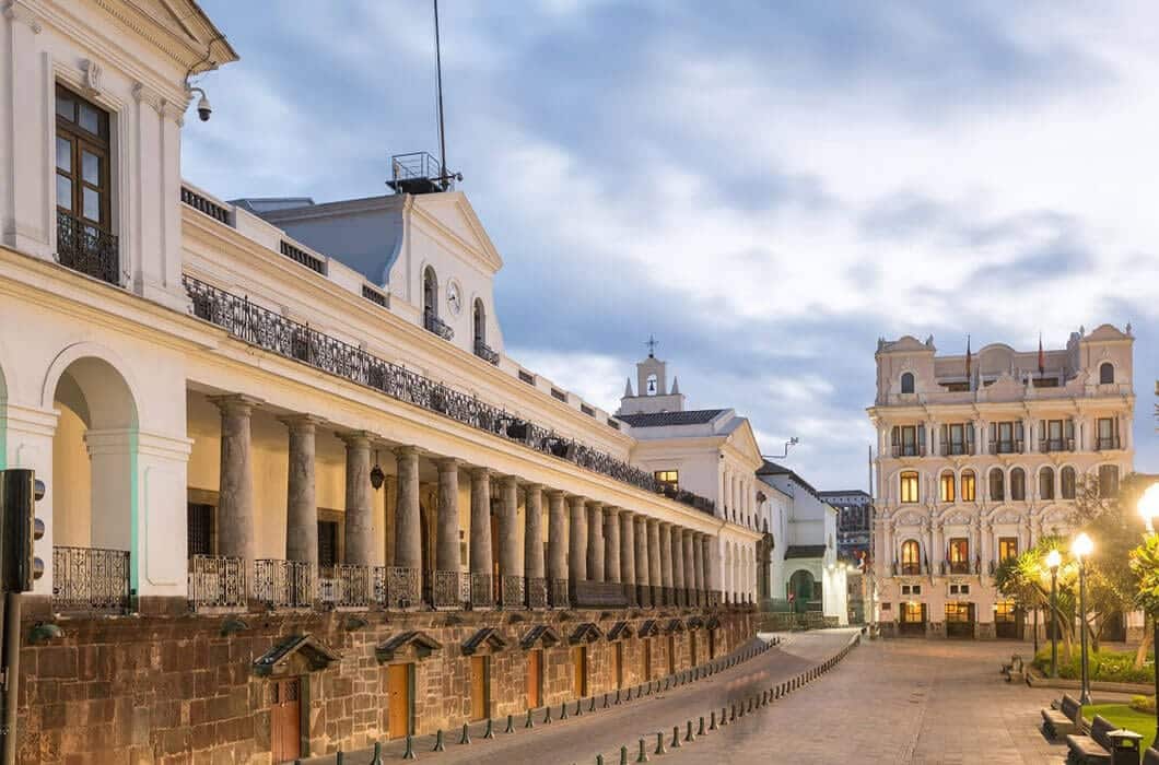 Colonial-style government buildings with columns and balconies line a cobblestone street at dusk, their facades warmly lit under a cloudy sky—an enchanting scene that highlights must-see Ecuador attractions.