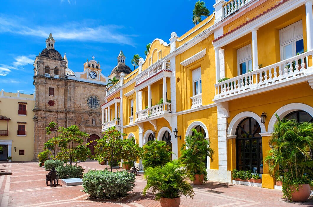 A vibrant yellow colonial building with white trim and balconies stands next to a historic stone church under a bright blue sky—a picturesque scene showcasing Colombia attractions in a sunny plaza dotted with potted plants and benches.
