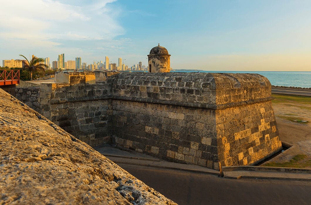 A stone fortification overlooks the sea, with a city skyline behind it under a clear sky. The old fort’s weathered walls contrast with modern buildings, making it one of the top Colombia attractions for travelers seeking history and views.