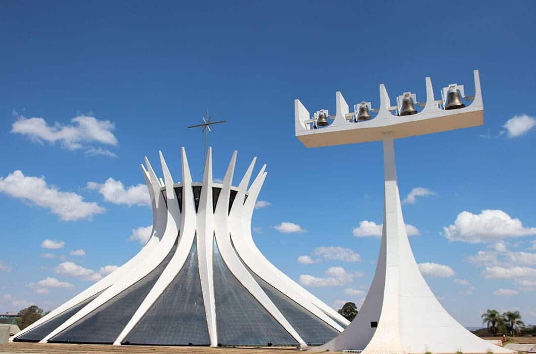 A modern white cathedral with curved columns and a cross on top stands beside a tall white bell tower under a blue sky—one of the striking Brazil attractions perfect for your Brazil travel itinerary.