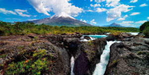 A river flows through dark volcanic rocks surrounded by lush greenery, with a snow-capped volcano rising in the background—an iconic scene among Chile attractions—under a bright blue sky with scattered clouds.