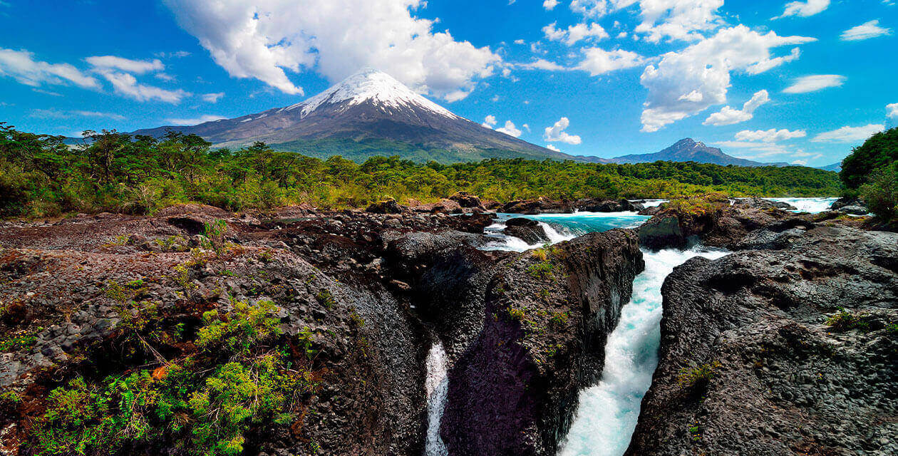 A river flows through dark volcanic rocks surrounded by lush greenery, with a snow-capped volcano rising in the background—an iconic scene among Chile attractions—under a bright blue sky with scattered clouds.