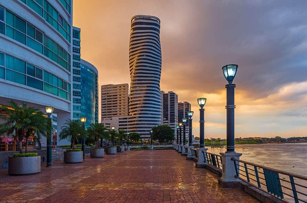 A riverside promenade at sunset, lined with modern high-rise buildings, palm trees, and street lamps—a must-see among Ecuador attractions. The golden light reflects on the river, creating a scenic highlight for Ecuador travel.