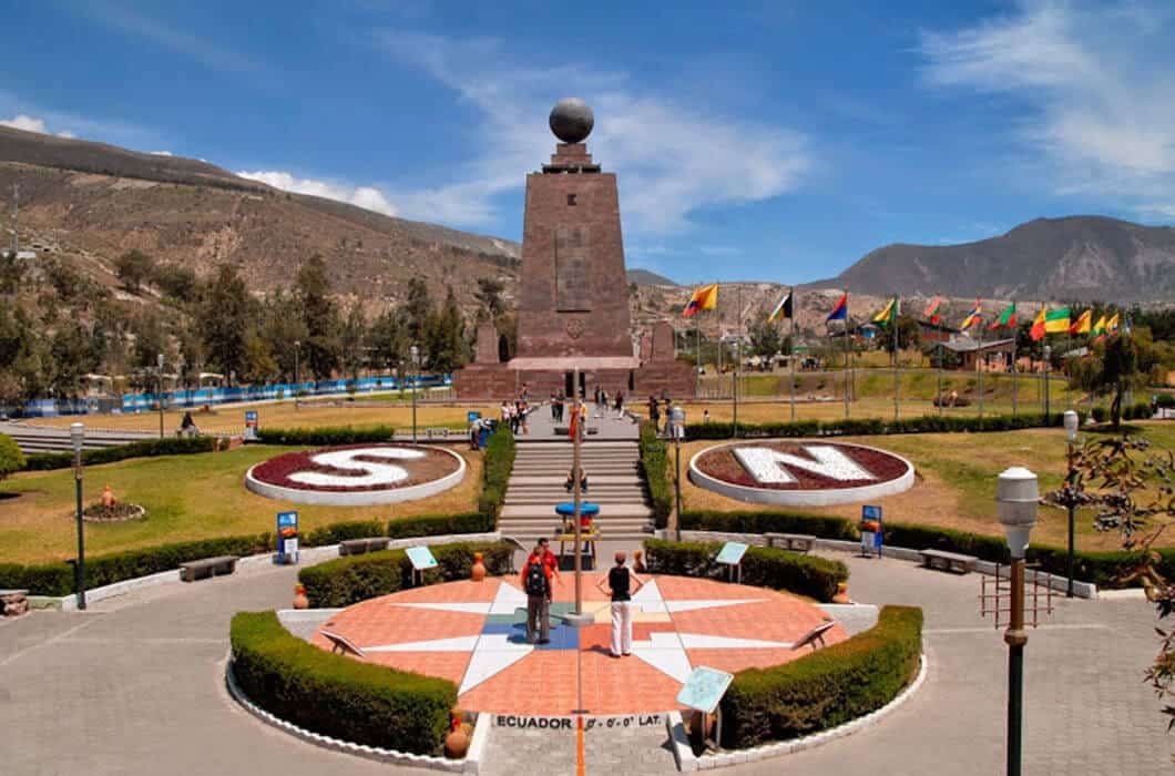 A monument with a large globe on top stands at the center of a plaza, surrounded by gardens and mountains. One of the top Ecuador attractions, it's a must-see for visitors exploring things to do in Ecuador along the equator line.