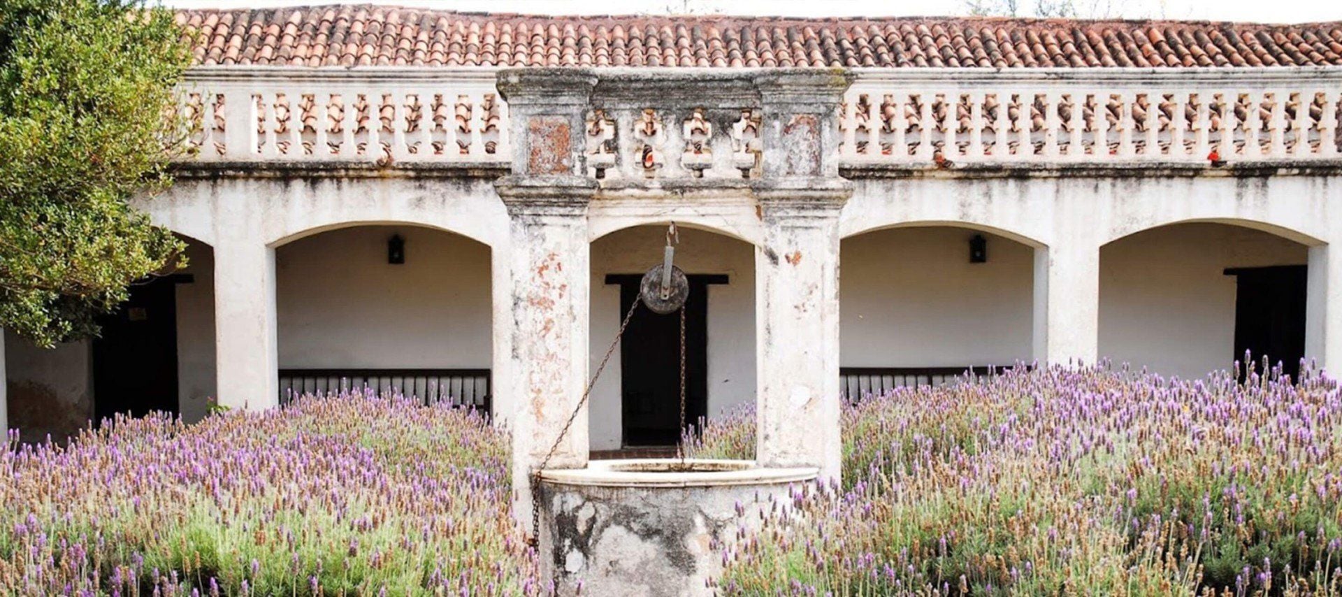 An old stone well stands in the middle of a courtyard filled with lavender, bordered by an aged, white, arched building with a tiled roof and decorative balcony—reminiscent of Cordoba's UNESCO World Heritage Jesuit Missions.