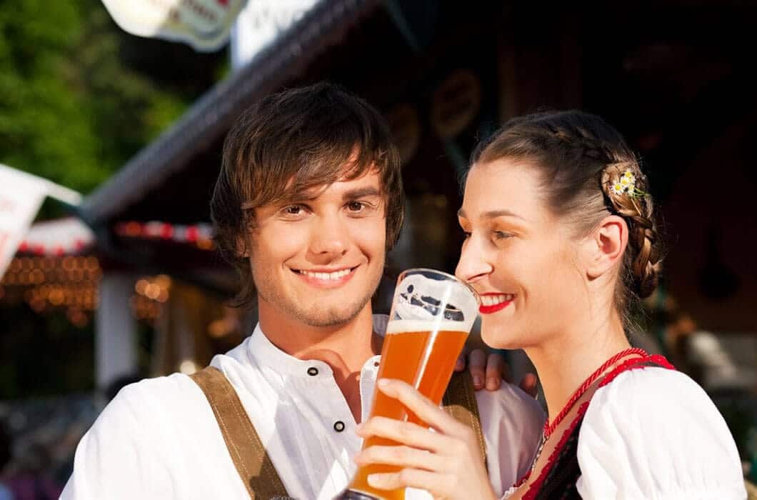 A smiling young man and woman in traditional Bavarian clothing enjoy a large glass of beer together outdoors, evoking the festive spirit you'd find among Brazil attractions—perfect for those seeking unique things to do in Brazil.
