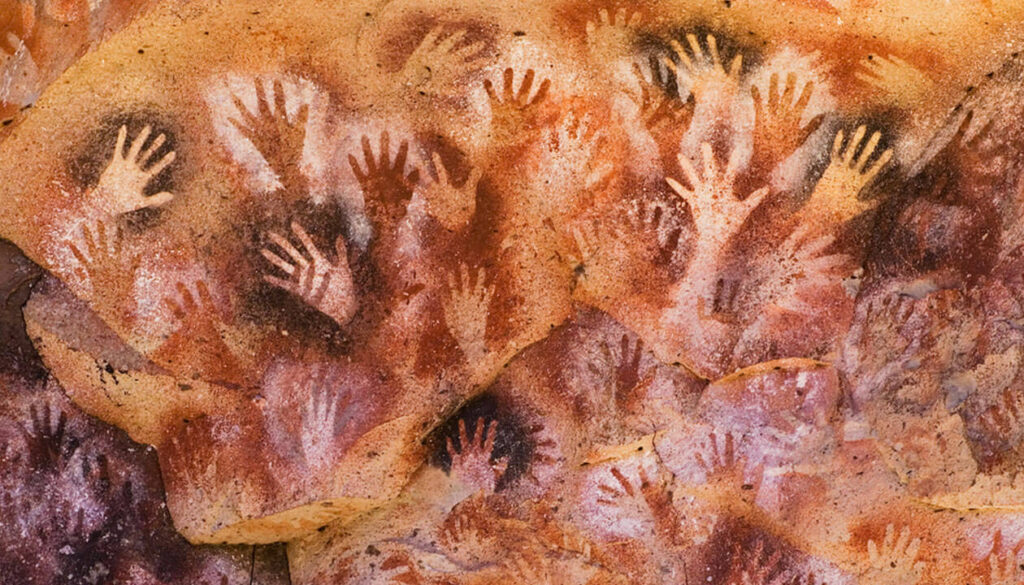 Hand stencils in various sizes cover a textured, reddish-brown stone wall, creating overlapping patterns typical of ancient cave art—discover the iconic Cave of the Hands in Argentina.