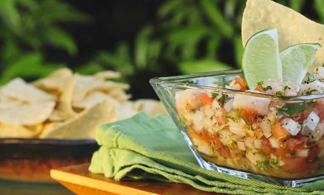 A glass bowl of ceviche garnished with lime wedges and tortilla chips sits on a green napkin, capturing the vibrant flavors that make trying local cuisine one of the top things to do in Peru. More tortilla chips are blurred in the background.