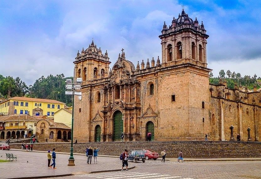 A large stone cathedral with ornate towers stands in a busy plaza, where people gather and relax. Colorful buildings and green trees surround the scene, capturing the festive spirit of celebrations in South America under a partly cloudy sky.