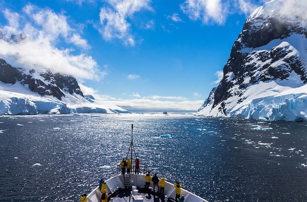 A group of people stand on the deck of a ship, sailing through icy waters between snow-covered mountains and glaciers—a breathtaking scene and one of the top Antarctica attractions under a bright blue sky with scattered clouds.