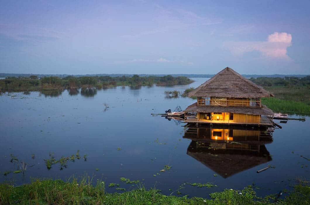 A thatched-roof house floats on a calm lake in Ecuador at dusk, its warm interior lights reflecting on the water. Surrounded by aquatic plants, this tranquil spot invites travelers to relax and discover unique things to do under a clear sky.