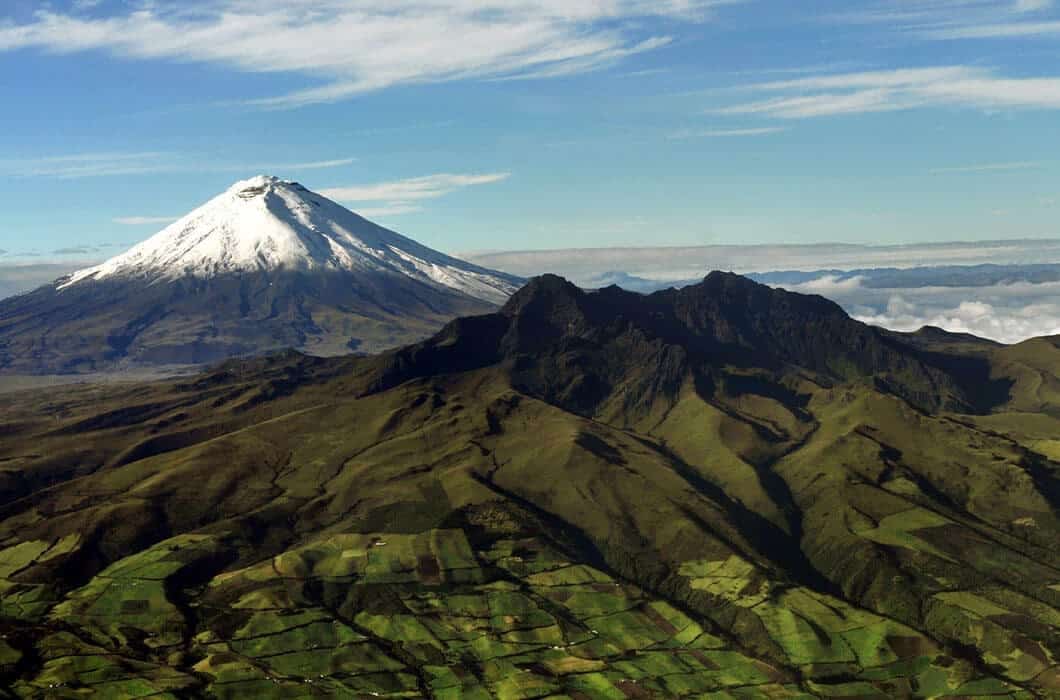 Snow-capped Cotopaxi volcano rises behind the green, rugged Rumiñahui mountains and patchwork farmland under a partly cloudy blue sky—one of the top Ecuador attractions for nature lovers.
