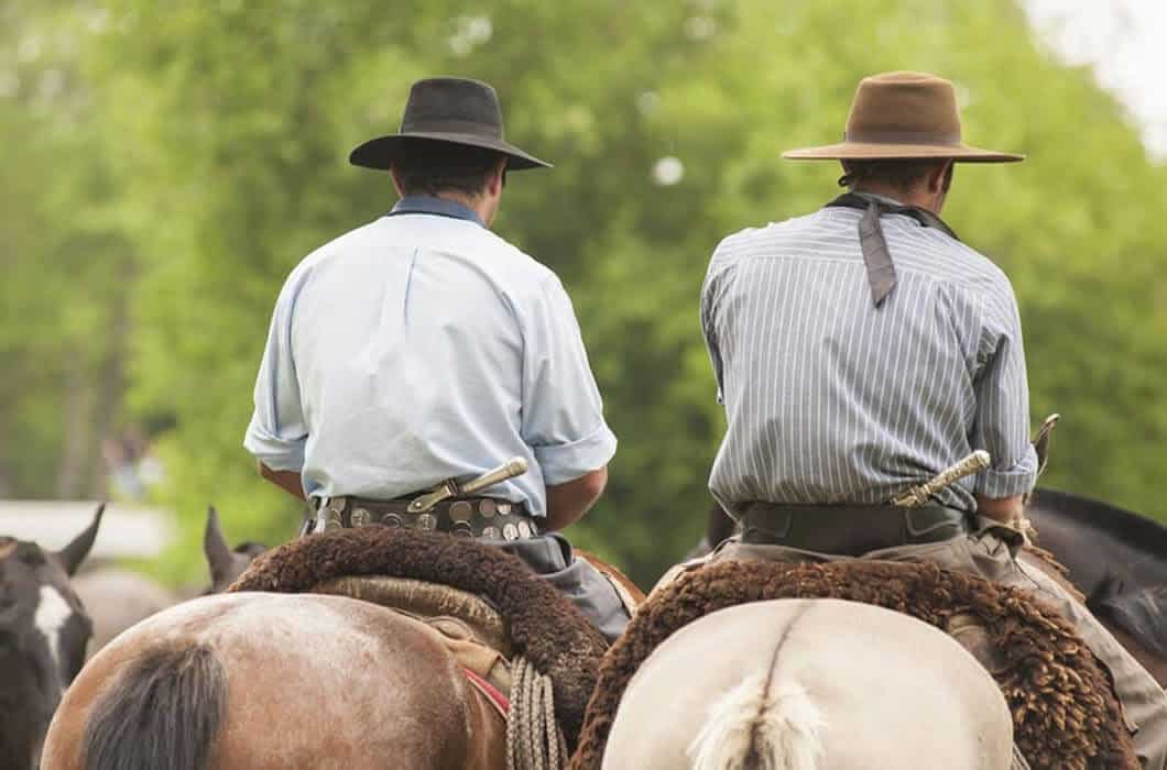 Two men wearing wide-brimmed hats and button-up shirts ride horses side by side, seen from behind, with green trees in the blurred background—an iconic travel experience and one of the top things to do in Patagonia.