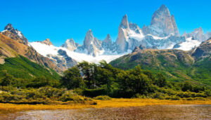 Sharp, jagged snow-capped mountains rise above green hills and dense forest, with a clear blue sky overhead and a calm river reflecting the vibrant landscape—one of the stunning places to visit on your Argentina travel adventure.