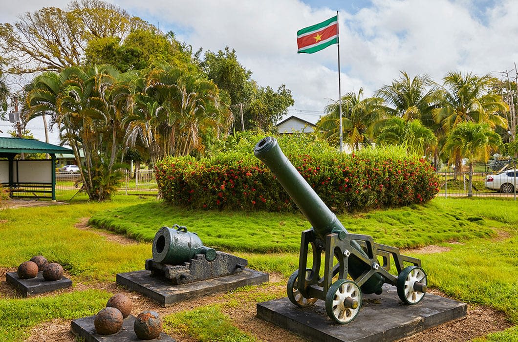 Historic cannons and cannonballs are displayed on grassy grounds with palm trees, and the Suriname flag flies on a tall flagpole—a must-see for those exploring Suriname or seeking things to do in Guyana and French Guiana nearby.
