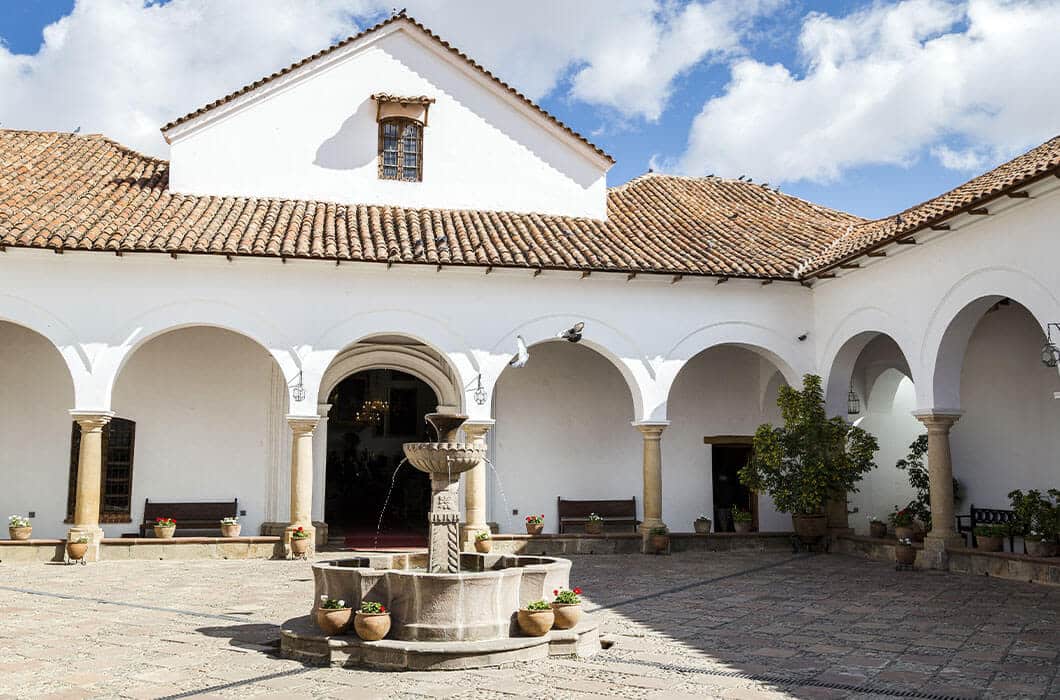 A courtyard with white arches, a tiled roof, and a central stone fountain surrounded by potted plants under a partly cloudy sky—one of the charming Bolivia attractions to discover as you explore things to do in Bolivia.