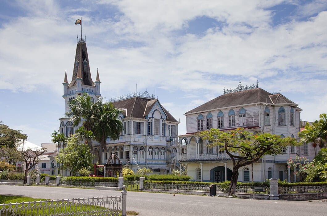 A large, ornate wooden building with steep roofs, a tall tower with a flag, arched windows, and decorative railings stands among palm trees—a must-see for those seeking things to do in Suriname under the bright blue sky.