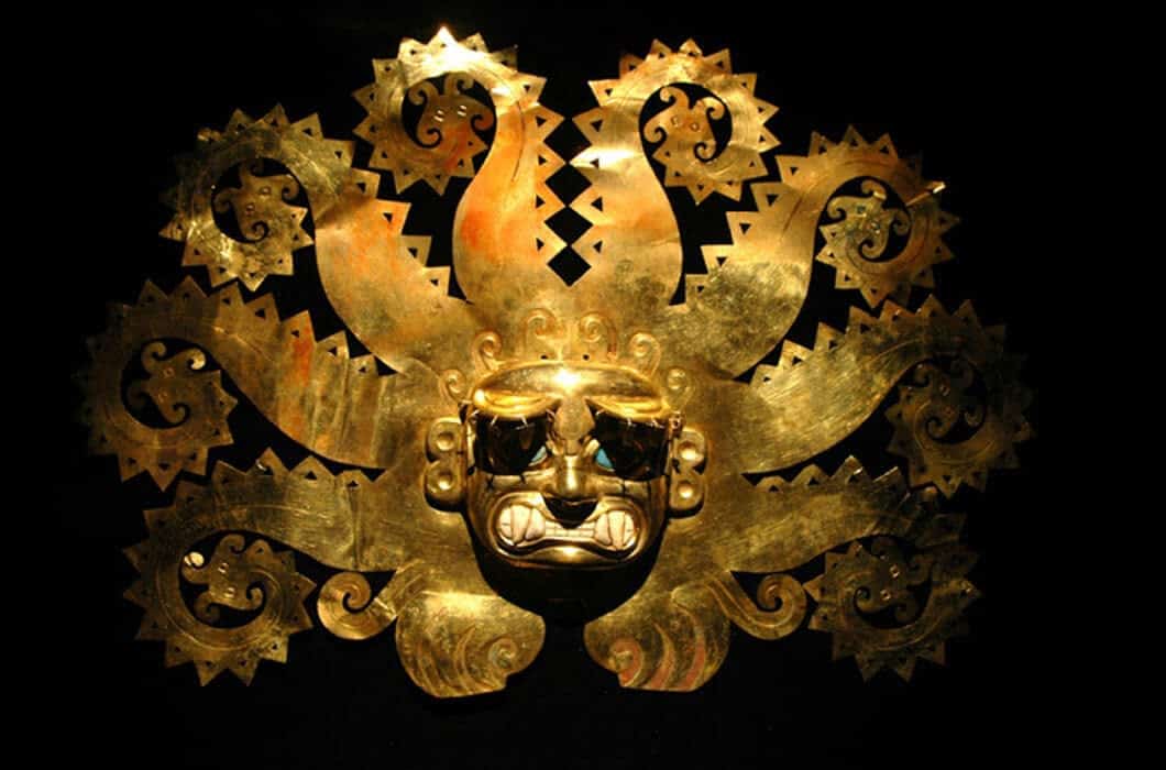 An ornate gold mask with a fierce face and multiple radiating, decorative rays or tentacles, displayed against a black background—a striking example of Colombia attractions for those interested in history and art.