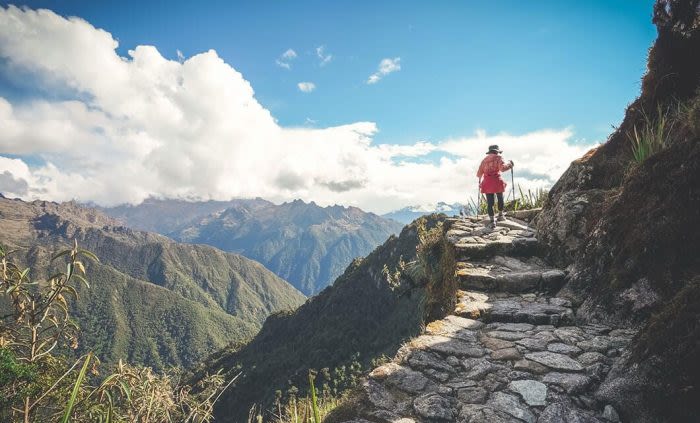 A person wearing a pink jacket and hat walks along a stone path on a mountain trail—one of the unforgettable Peru attractions—surrounded by green hills and distant peaks under a partly cloudy blue sky.