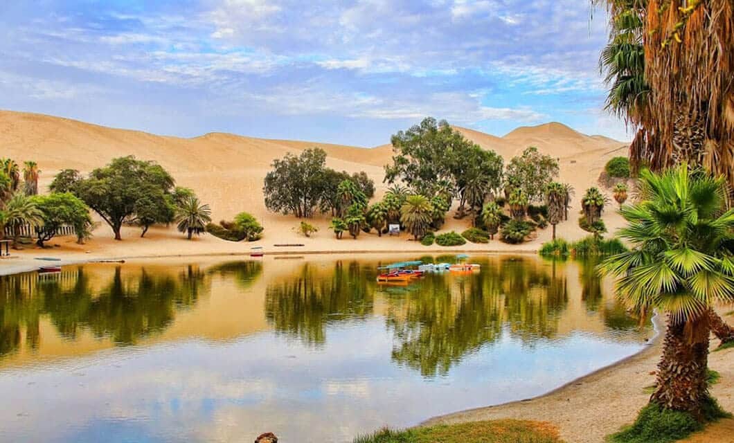 A calm oasis with palm trees and greenery surrounds a small lake, colorful paddle boats float on the water—one of the unique Peru attractions you’ll find amid sandy desert dunes under a partly cloudy sky.
