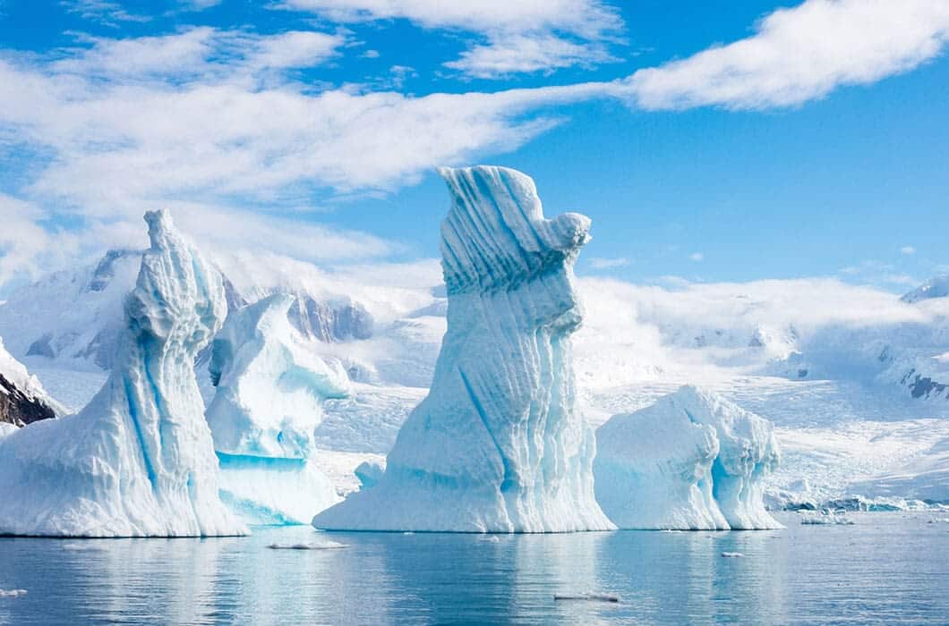 Large, jagged icebergs float in calm, blue water with snowy mountains and a bright, partly cloudy sky in the background—a breathtaking view often admired on Antarctica tours.