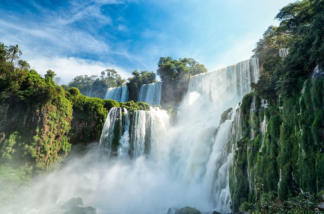 A powerful, multi-tiered waterfall cascades over lush, green cliffs surrounded by dense forest under a blue sky with scattered clouds—one of the stunning Paraguay attractions for nature lovers and a must-see highlight on any Paraguay tourism adventure.