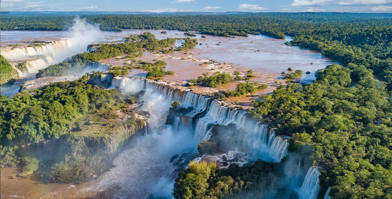 Aerial view of Iguazu Falls with wide, powerful waterfalls cascading over cliffs, surrounded by lush green forest and mist rising from the water below.