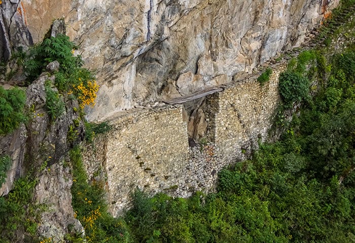 A narrow stone path clings to a steep cliffside near Machu Picchu, with a section missing above dense green foliage. The rocky wall and overgrowth highlight the trail’s rugged and precarious nature—a must-see for adventurous travel.