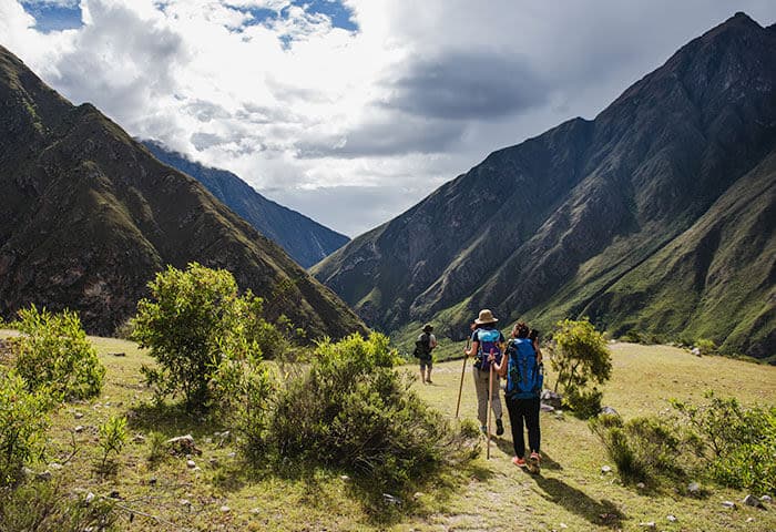Three hikers walk along a grassy path between green mountain slopes under a mostly cloudy sky, surrounded by shrubs and dramatic peaks—an inspiring glimpse into South America travel and its breathtaking attractions.