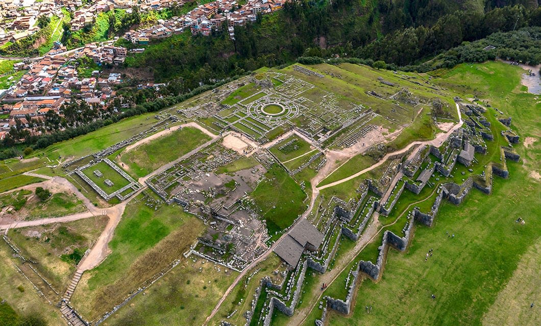 Aerial view of the ancient stone ruins of Sacsayhuamán in Peru, with zigzagging walls and grassy areas—a must-see among Peru attractions—overlooking a nearby town surrounded by forested hills.