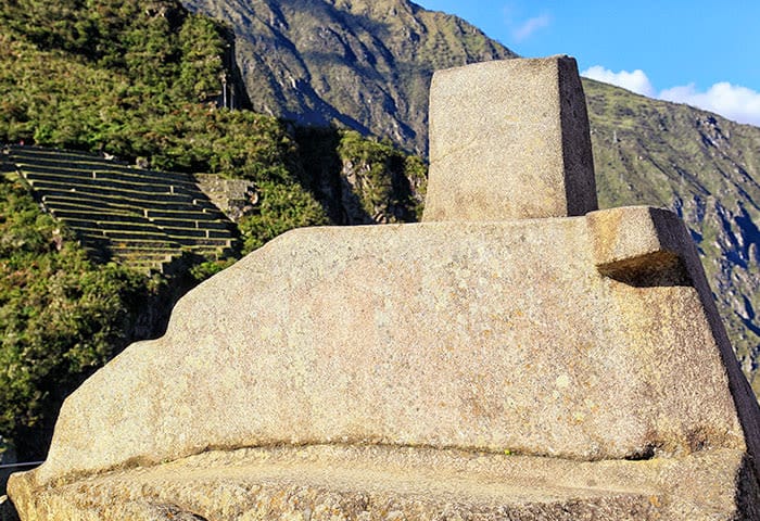 A close-up of the Intihuatana stone at Machu Picchu, Peru—one of the top things to do when you travel here—set against terraced green hills and majestic mountains beneath a clear blue sky.