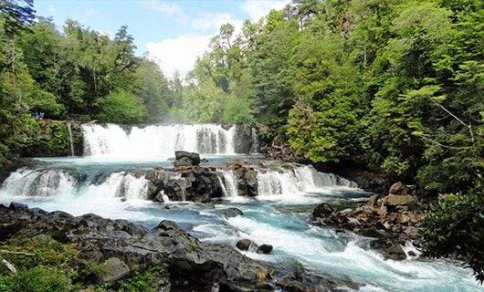 A wide waterfall cascades over dark rocks in the Huilo-Huilo Reserve, surrounded by dense green forest under a partly cloudy sky. The water flows into a clear, blue stream with rocky edges in the foreground.
