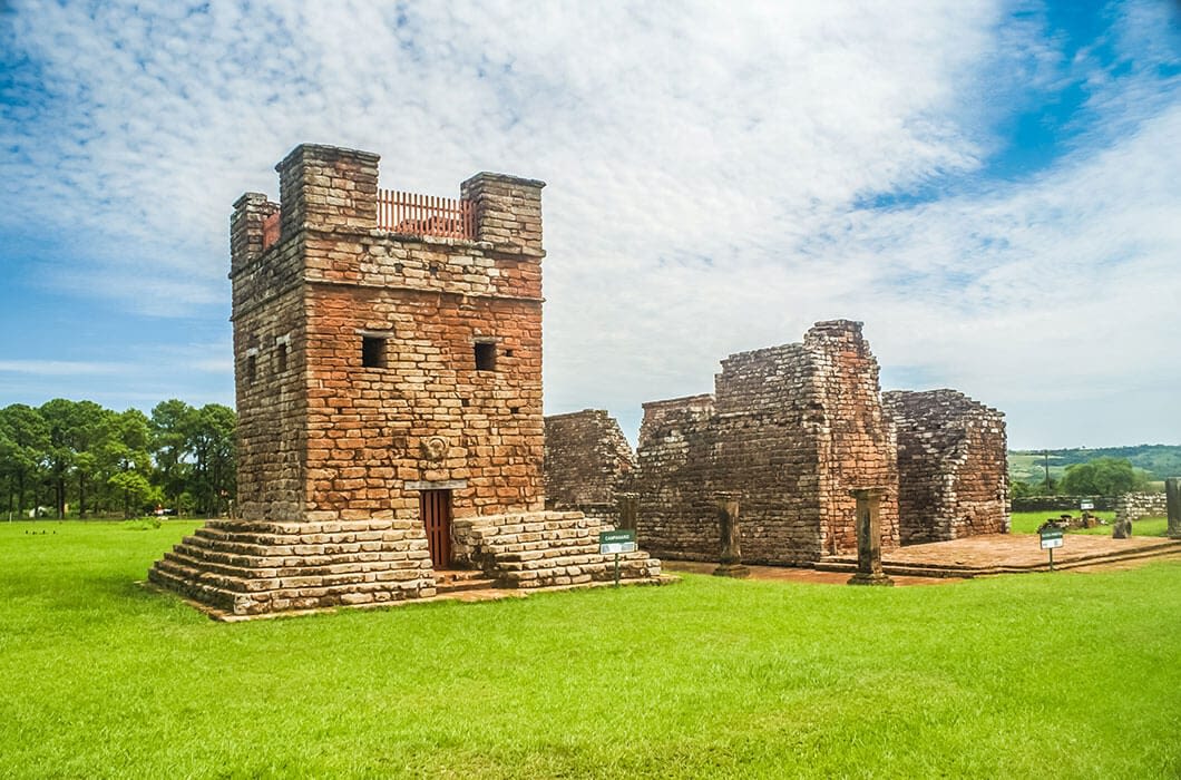 Ancient brick ruins of a Jesuit mission stand on a grassy field under a partly cloudy sky, with a tall square tower and partially collapsed walls—one of the top Paraguay attractions for those seeking unique things to do in Paraguay.