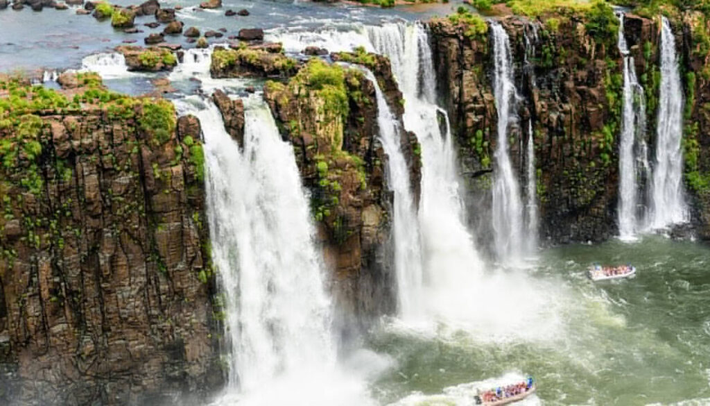 Large, powerful waterfalls cascade over rocky cliffs surrounded by lush greenery. Tour boats with passengers navigate the river below, creating mist and a dramatic scene—truly one of South America's Best Places to Visit, as captured by Juergen Keller.