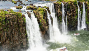Large, powerful waterfalls cascade over rocky cliffs surrounded by lush greenery. Tour boats with passengers navigate the river below, creating mist and a dramatic scene—truly one of South America's Best Places to Visit, as captured by Juergen Keller.