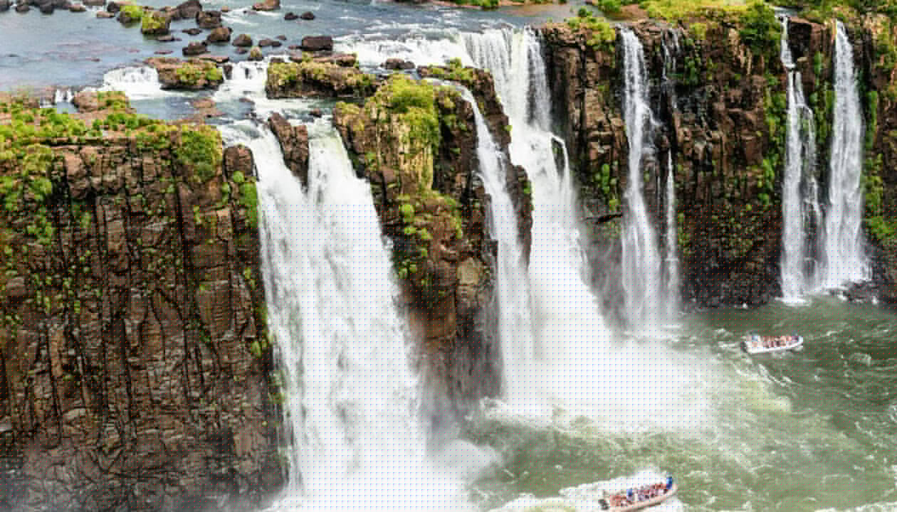 Large, powerful waterfalls cascade over rocky cliffs surrounded by lush greenery. Tour boats with passengers navigate the river below, creating mist and a dramatic scene—truly one of South America's Best Places to Visit, as captured by Juergen Keller.