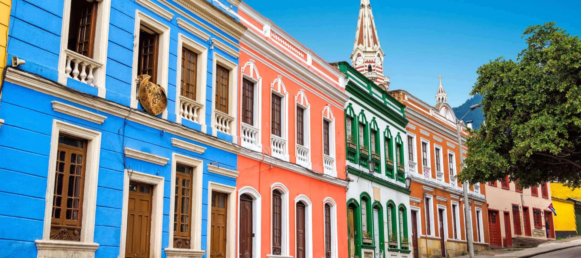 Row of colorful colonial-style buildings in blue, orange, green, and white with decorative windows and balconies, under a sunny sky. Perfect for exploring on Bogota Tours, with a leafy tree adding charm to the vibrant scene.