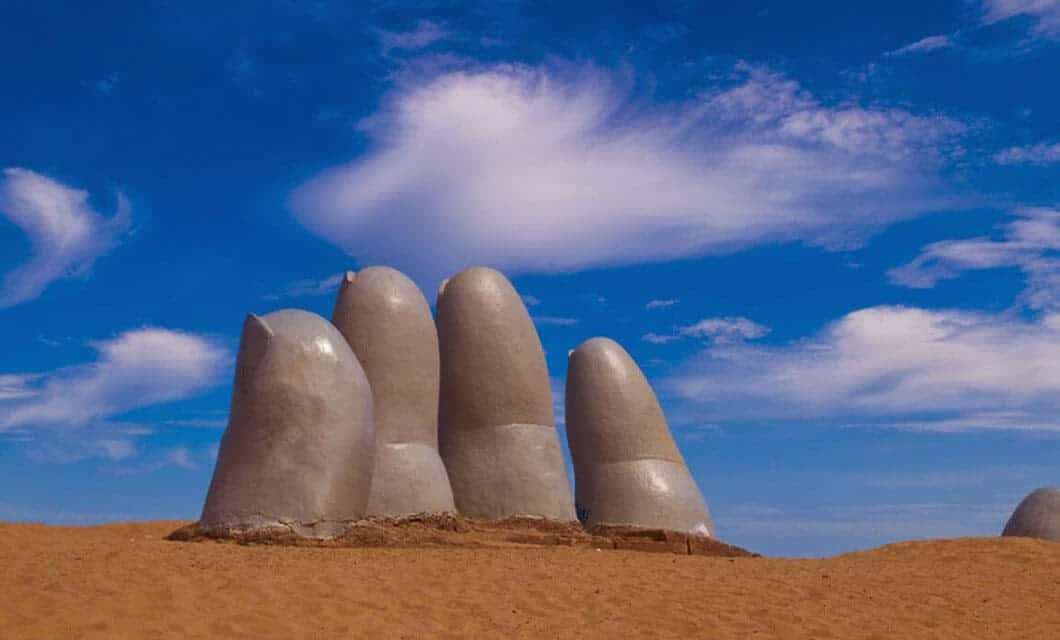 A giant hand sculpture with four fingers and a thumb emerges from sandy ground under a bright blue sky with wispy clouds—one of the most iconic Uruguay attractions for anyone interested in unique things to do in Uruguay.