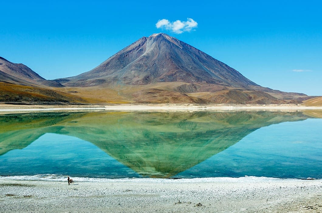 A tall, cone-shaped mountain with a small cloud above it is reflected in a clear, turquoise lake—one of the stunning Bolivia attractions. Dry, brown terrain surrounds the lake as a person stands near the water’s edge in the lower left corner.