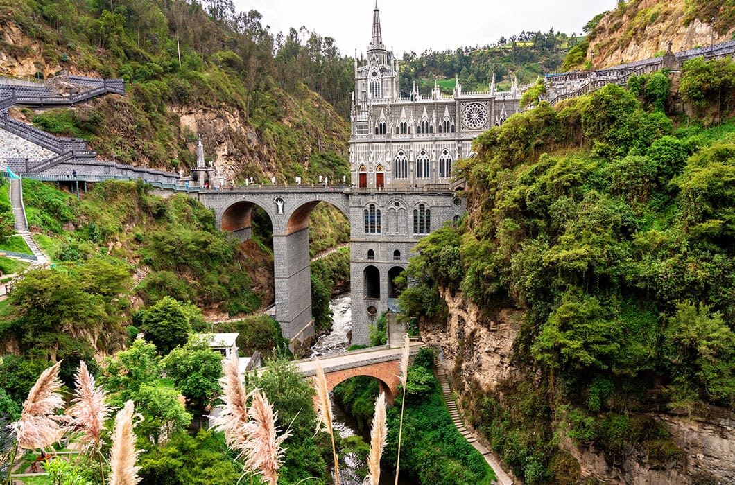 A striking Gothic-style church, Las Lajas Sanctuary, sits atop a stone bridge spanning a deep green canyon—a must-see among Colombia attractions, surrounded by lush hills and vegetation in southern Colombia.