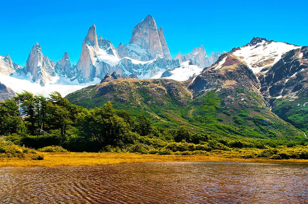 Snow-capped jagged mountain peaks rise above lush green hills and a clear river under a bright blue sky in Patagonia. Dense trees cover the lower slopes, making it a perfect travel destination with endless things to do and explore.