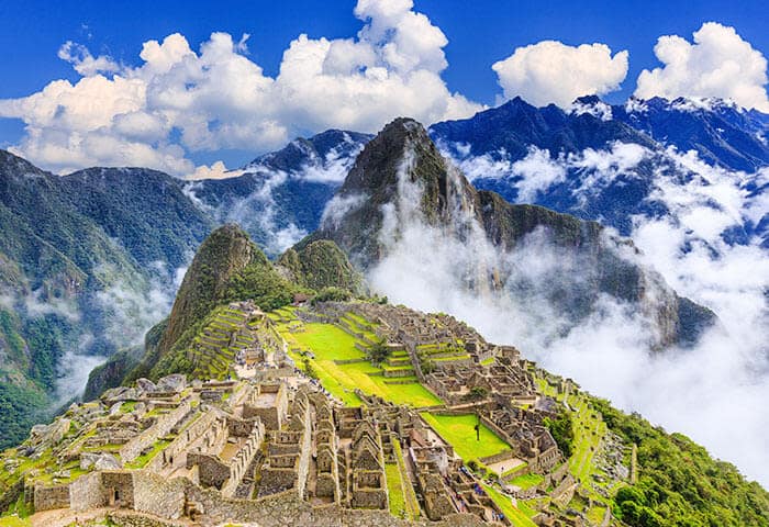 Ancient stone ruins of Machu Picchu sit atop a lush green mountain ridge, surrounded by mist and dramatic peaks—one of the most breathtaking things to do under a bright blue sky with scattered white clouds.