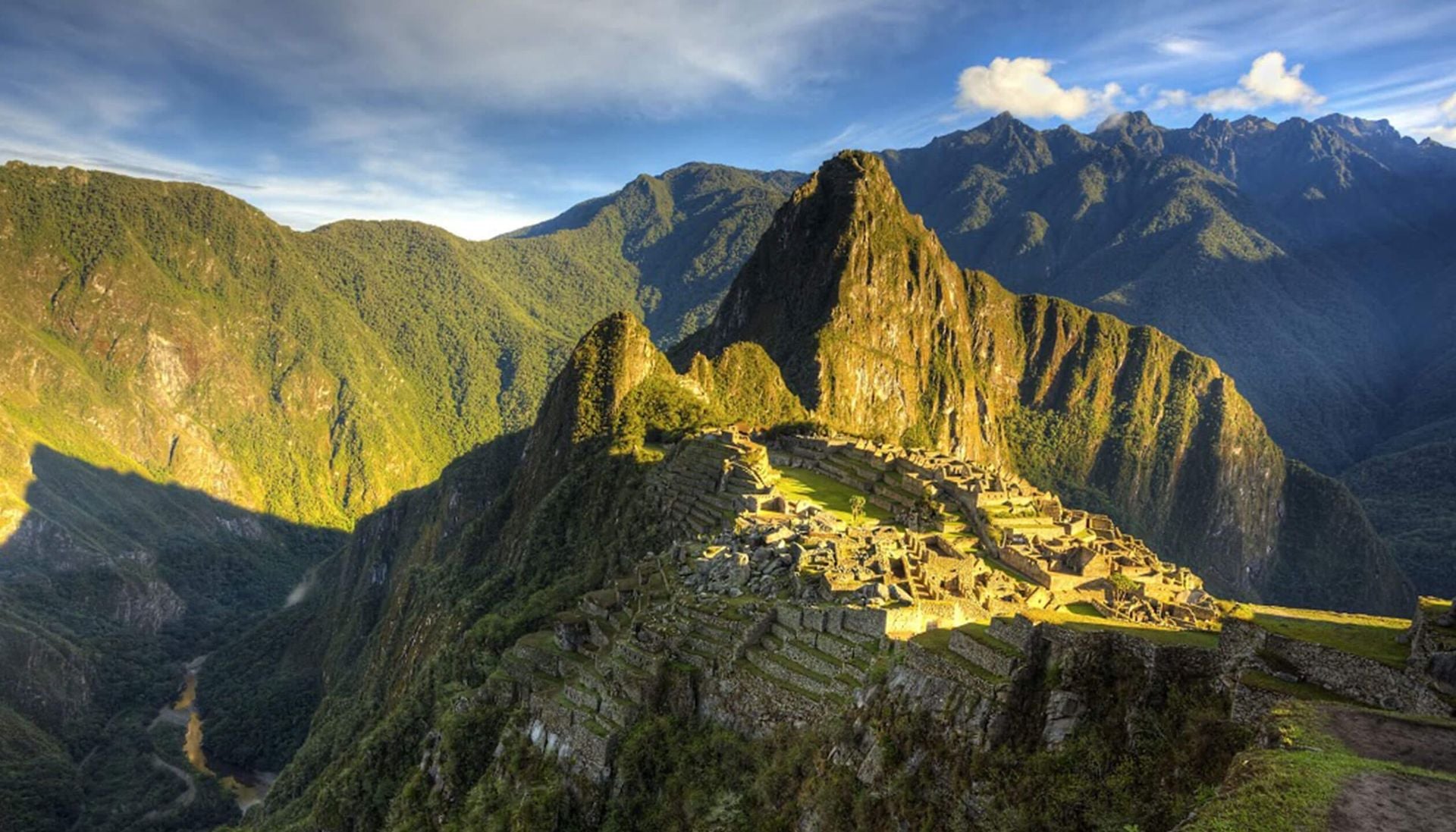 Ancient stone ruins of Machu Picchu sit atop a lush mountain ridge, surrounded by green peaks and bathed in golden sunlight, as the echoes of traditional Peruvian music drift through the partly cloudy blue sky.