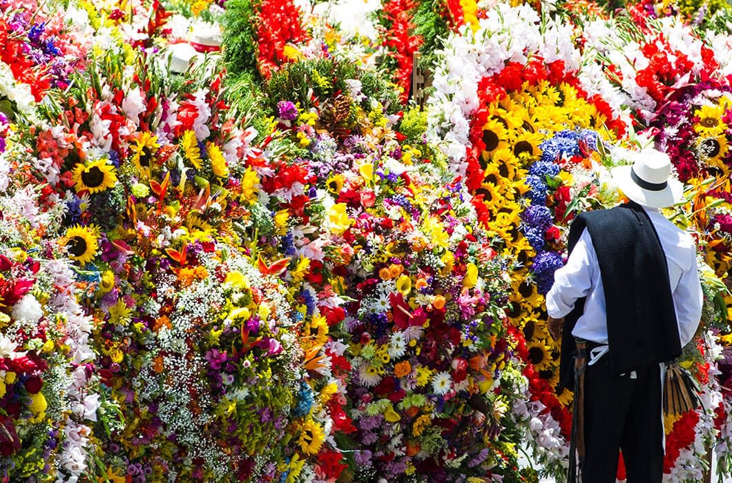 A person wearing a white hat and dark clothing stands in front of large, colorful flower arrangements at an outdoor festival or parade—an experience often featured on Medellin Tours. Bright sunflowers, lilies, and various blooms fill the scene.