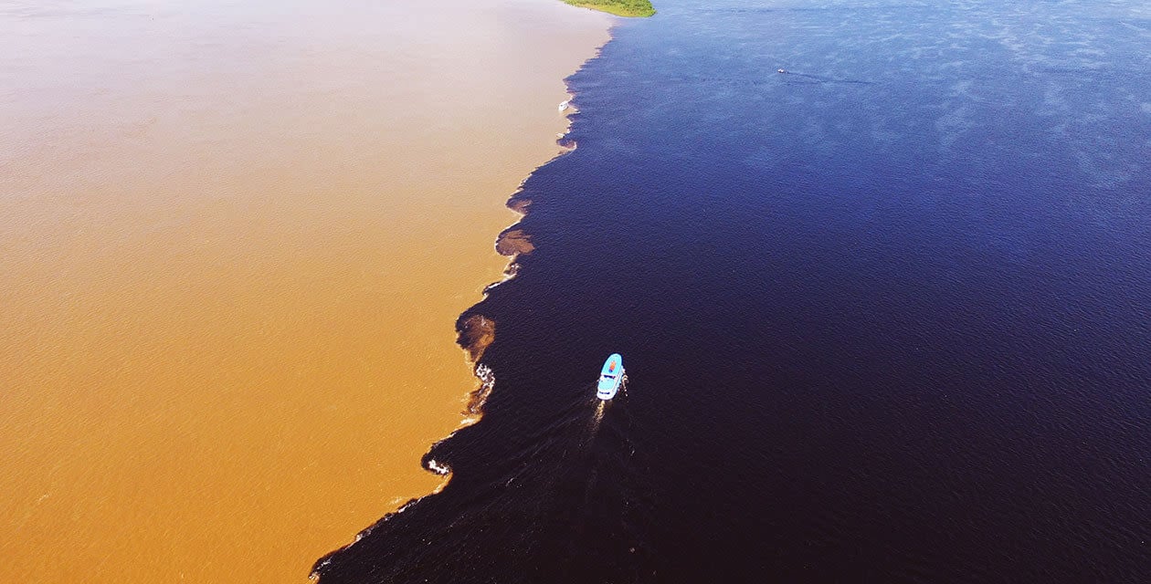 A small boat travels along the line where muddy brown water meets dark blue water on an Amazon Tour from Manaus, showing a sharp contrast between the two, with a small patch of green land in the distance.