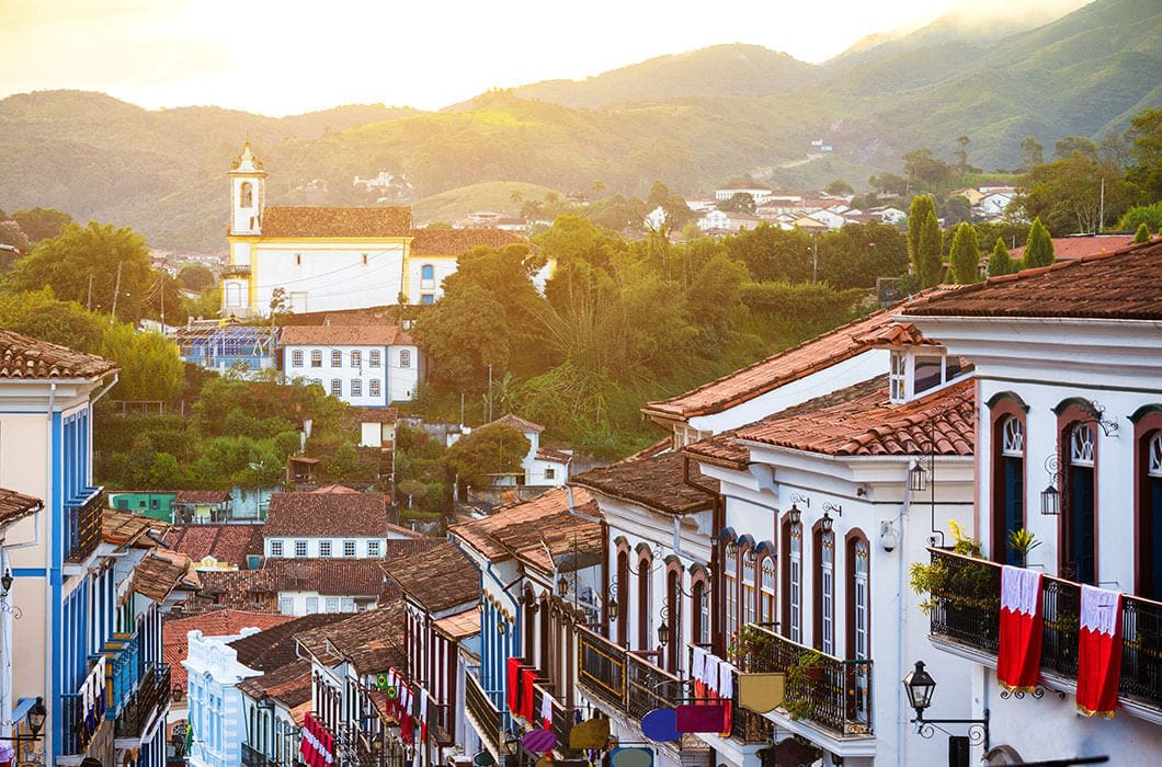 Colorful colonial houses line a steep cobblestone street, laundry draped from balconies, leading toward a white church and green hills—capturing the charm of Brazil tourism beneath a glowing sunset sky.