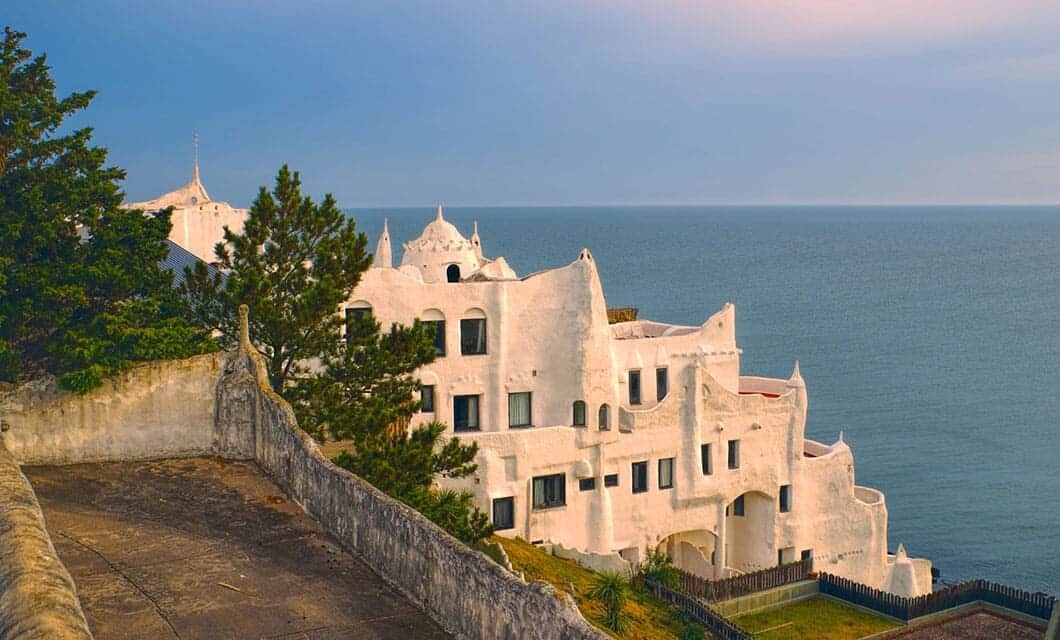 White, Mediterranean-style building with rounded edges and unique architecture sits on a hill overlooking the blue ocean—one of the must-see Uruguay attractions—with trees and a pathway in the foreground under a soft, pastel sky.