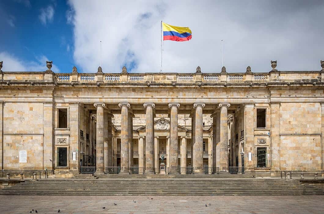 A large neoclassical building with columns, stone steps, and the Colombian flag flying on top. The sky is partly cloudy, and several pigeons are on the plaza in front—one of the top Colombia attractions for visitors.