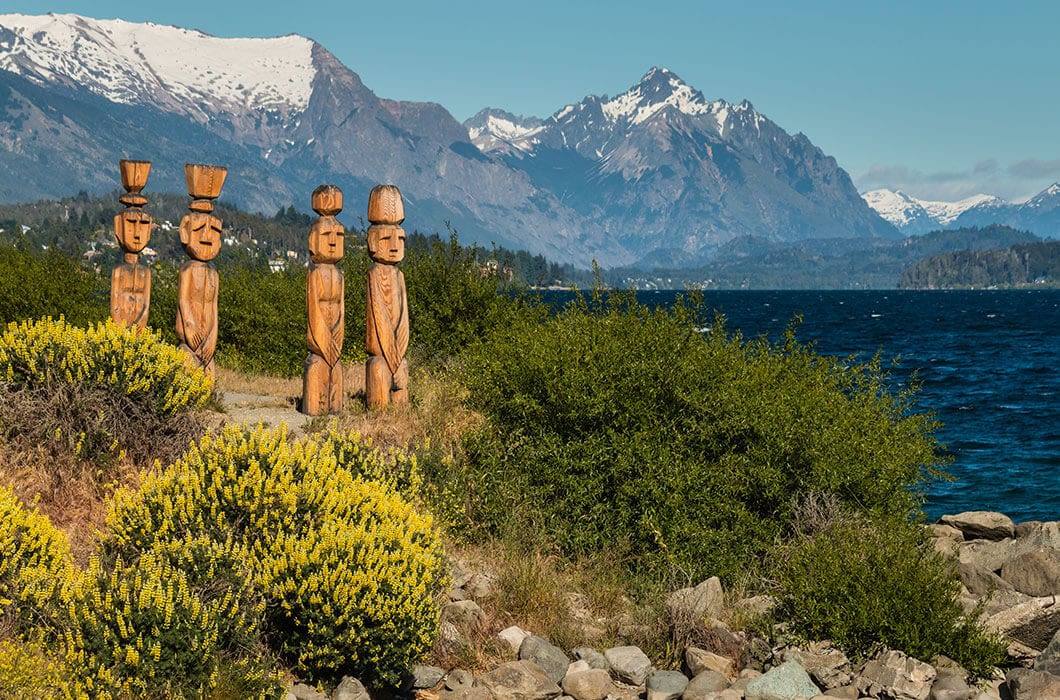 Wooden totem statues stand by a lakeshore in Patagonia, surrounded by green bushes and yellow flowers, with snow-capped mountains and blue sky in the background—an inspiring spot for travel and things to do.