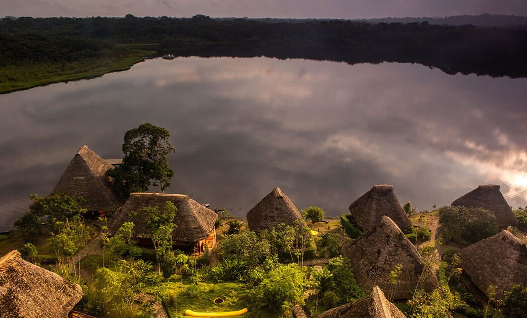 Aerial view of thatched-roof huts at Napo Wildlife Center Eco-Lodge, surrounded by lush greenery near the calm lake’s edge, set deep in the Ecuador Amazon with trees and forest in the background under a cloudy sky.